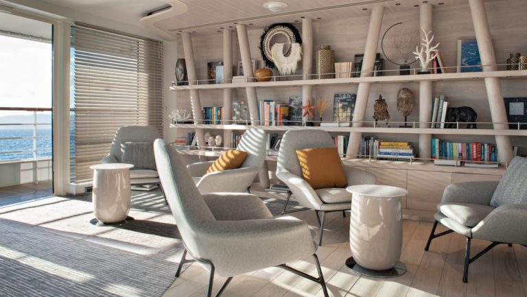 Library on Le Bougainville ship with padded armchairs, pedestal coffee tables, beige accents & bookshelf with ethnic art & books.