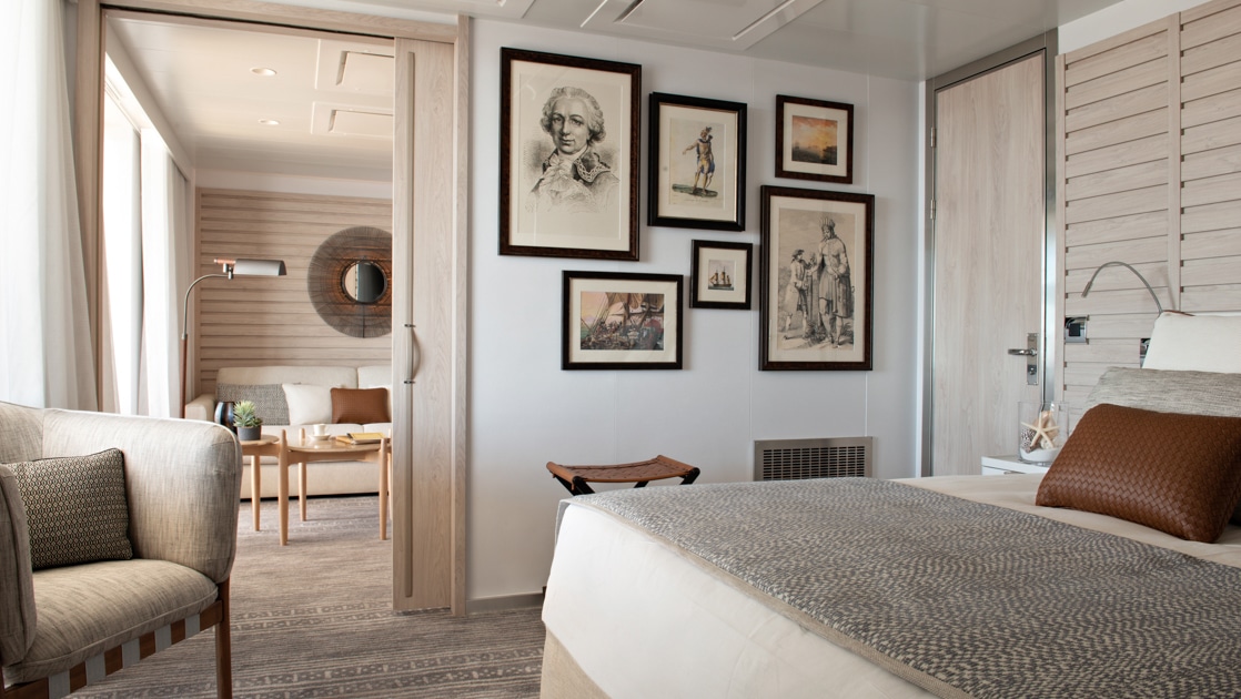 View from bedroom to window-lined living room with couch in the beige-accented Owner's Suite on Le Bougainville cruise ship.