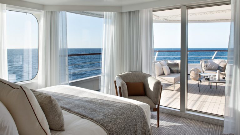 View from bed to outdoor terrace with wraparound windows in the beige-accented Owner's Suite on Le Bougainville cruise ship.