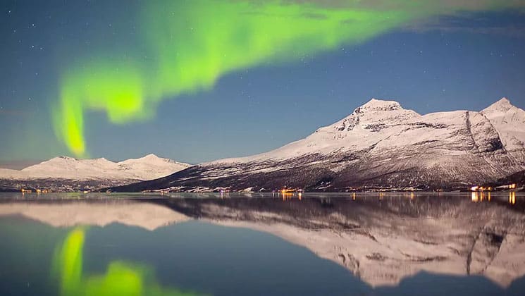 Green glow of Northern Lights spreads above a waterside town backed by snow-covered mountains reflecting in the glassy fjord.