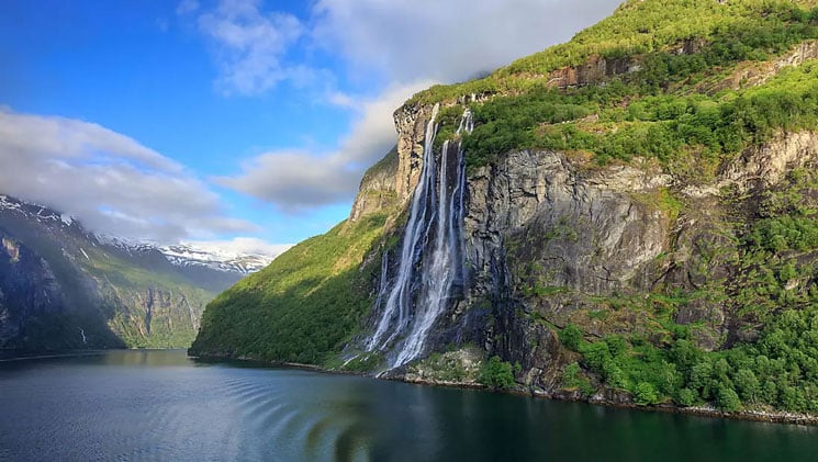 Large waterfall with 2 main falls spews over cliff face into glassy water by green hillsides on a Norwegian fjords cruise.