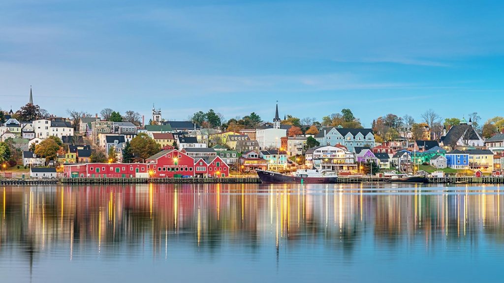 Hillside harbor town at dawn with docked fishing boat, colorful homes, church steeples, fall foliage & glassy water.