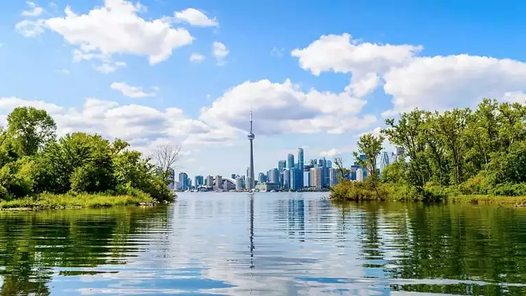 Toronto's CN Tower rises above a glassy river, green forested riverbanks & city high rises, seen on small ship great lakes cruises.