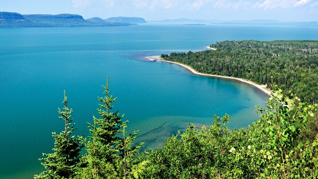 Aerial view looking down on Lake Superior on a calm day with its turquoise water, white-sand shoreline & bright green forest.