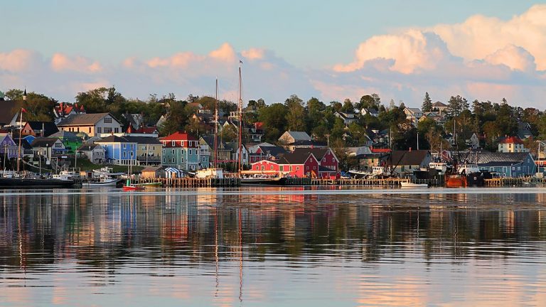 Hillside harbor town at dusk, seen on St. Lawrence River cruises with various boats, colorful homes, green trees & calm water.