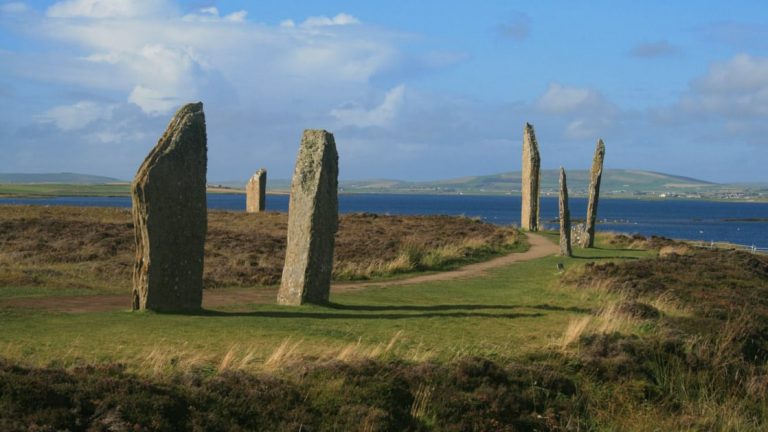 6 pillars of the Ring of Brodgar on the Orkney Islands are seen by a path through grass with the ocean and land behind them.