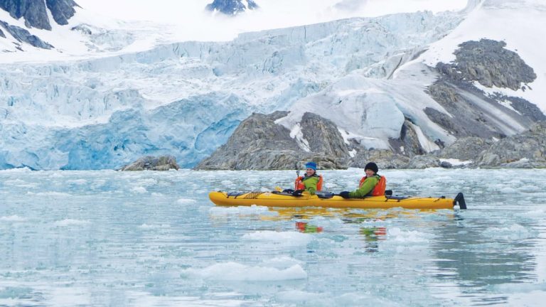 Two travelers in a yellow kayak seen still and smiling in icy Arctic water in front of a glacier.