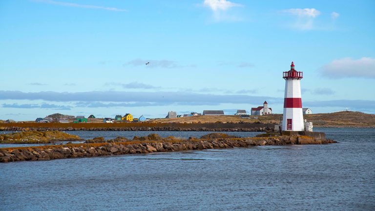 Red & white lighthouse sits on rocky sea wall by calm ocean under blue skies seen on a St. Lawrence river cruise.