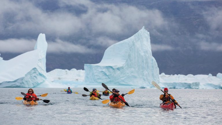 Five kayakers seen in the water in front of giant white icebergs.