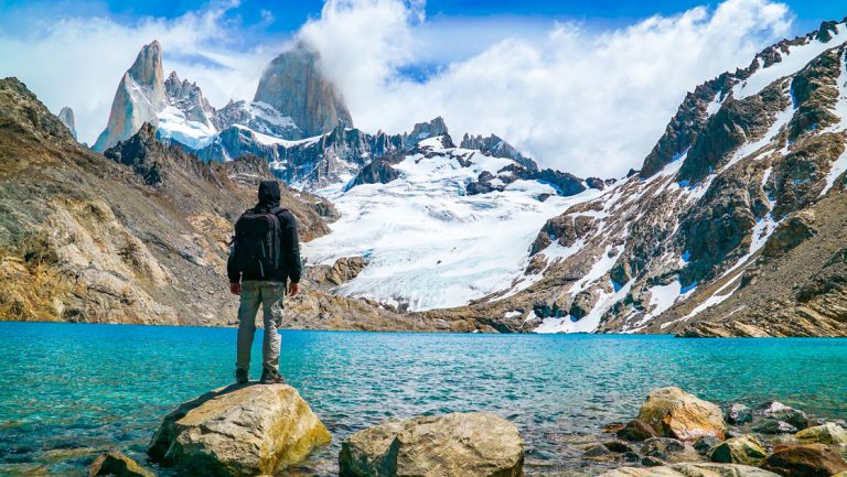 Explora El Chalten traveler stands on a boulder by a turquoise lake with a hanging snowfield under jagged peaks in the distance.