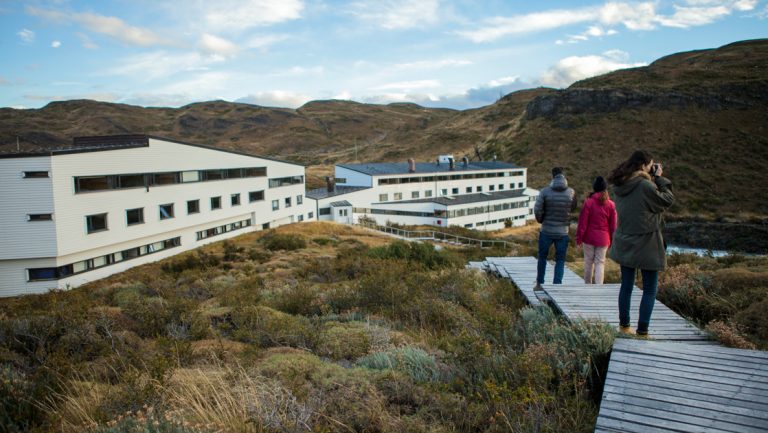Explora Connects travelers on a boardwalk by rectangular white hotel among pampas grassland in Torres del Paine National Park.