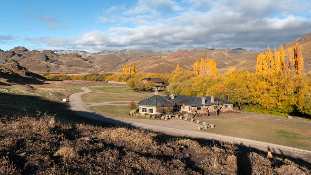 Aerial view of small stone wilderness lodge sitting in Patagonia National Park valley with yellow-leaf trees under some clouds.