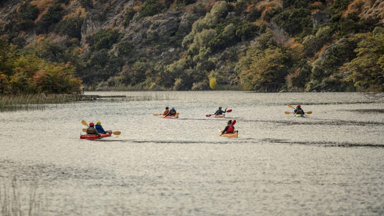 8 Explora Patagonia National Park kayakers paddle a wide, calm river beside hills of dense fall foliage & cliffs.