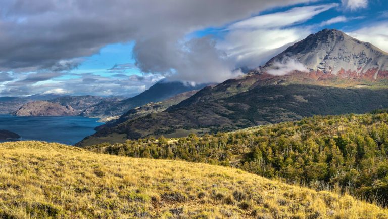 Landscape of grassy hills & volcanic mountains beside a bright blue lake under a partly cloudy sky in Patagonia National Park.