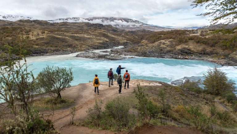 4 Explora Patagonia National Park travelers stand beside a large turquoise river as a guide points out flora & fauna.