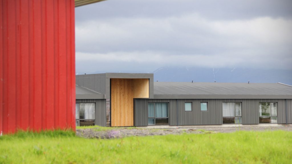 Red metal building & bright green grass front a modern gray building with wooden accent wall on a cloudy day in Iceland.