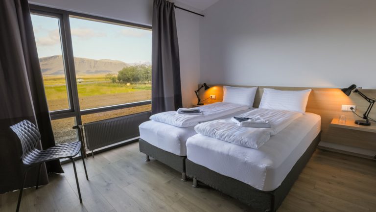 2 single beds pushed together with white linens by large window looking onto grassy field at Lilja Guesthouse in Iceland.
