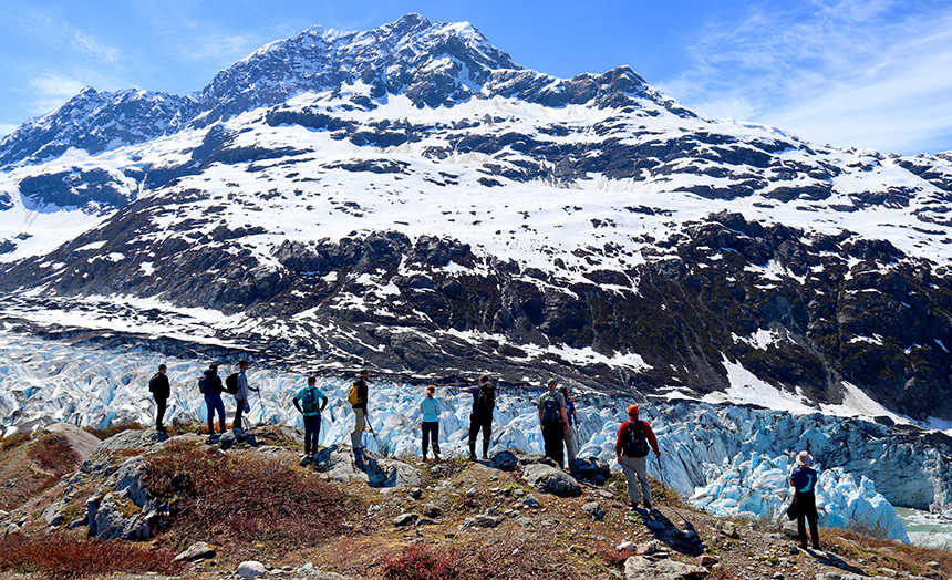 Alaska guests stand on a ridge overlooking a white and teal icy glacier flanked by a snowy mountain range as part of a daily cruise activity option. 