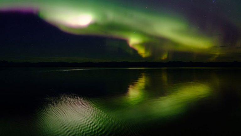 Northern lights in Alaska with green, yellow & purple plumes of light over glassy water & dark shadows of nearby mountains.