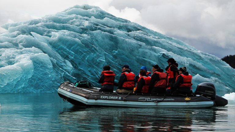 Black Zodiac inflatable boat with travelers in red life jackets sits beside a large, dark-blue iceberg on an Alaska winter cruise.