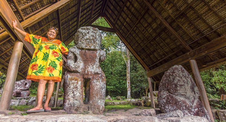 Woman in tropical-print dress stands beside stone statues under a thatch roof during an Ancient Polynesia small ship cruise.