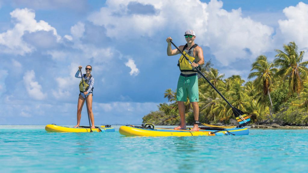 Young man & woman each paddle a yellow paddleboard in turquoise water with palm trees behind on an Ancient Polynesia cruise.