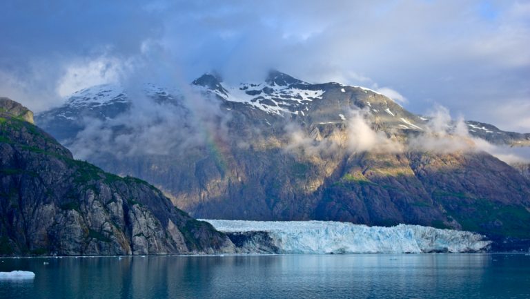 A large glacier meets the sea with rocky, snow-spotted mountains behind on a cloudy day in Glacier Bay National Park.