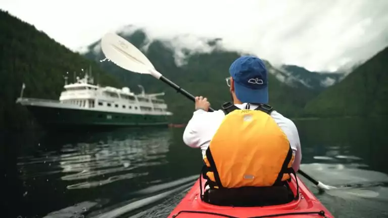 Man paddles a red kayak toward a small ship with dark green hull & white upper decks backed by forested hills.