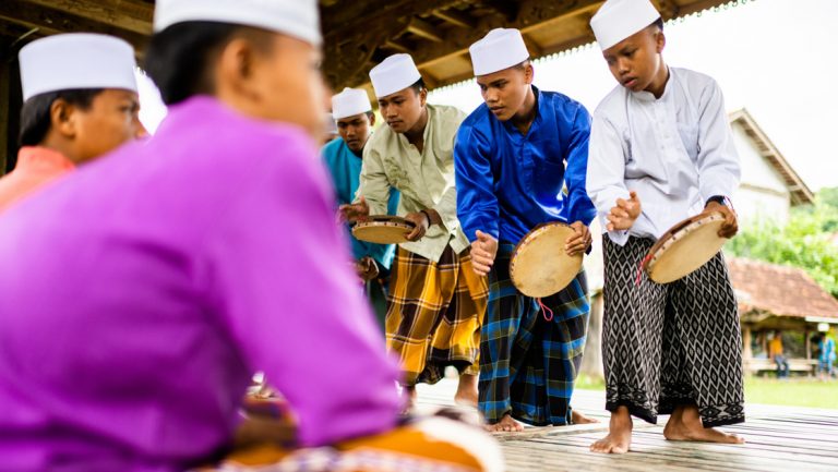 Indonesian boys & men in round white hats & multicolored, loose clothes sit & stand in lines while holding tambourines.
