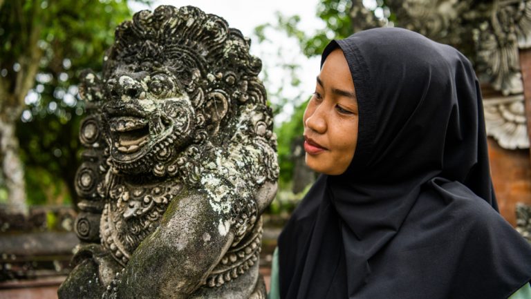 Woman in black head scarf looks at weathered stone lion statue at religious site of Lingsar in Lombok, Indonesia.