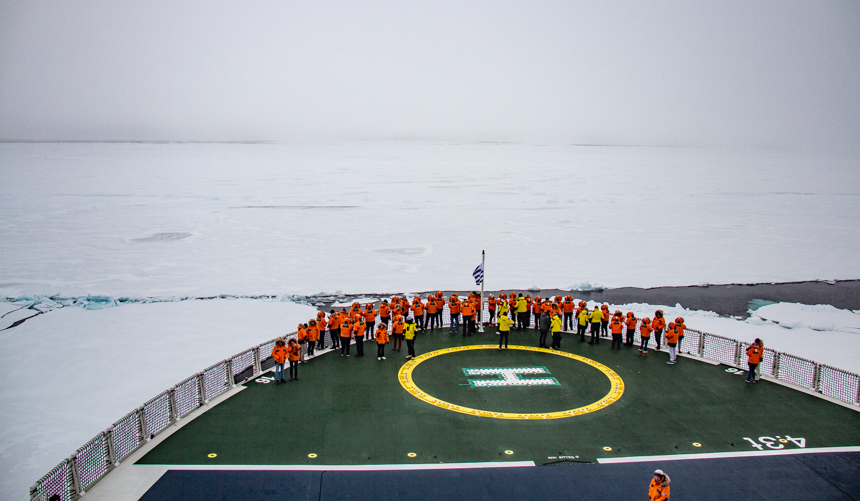 Guests in orange and yellow parkas gather on the bow of Charcot ship to view the all white expanse of sea ice at North Pole