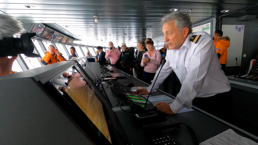 Captain of Le Commandant Charcot wears white collared shirt with striped epaulette and stands over control board in the bridge.