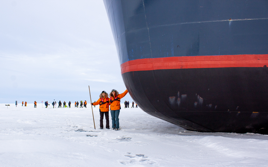 Two females in orange parkas pose touching the black red and blue painted hull of Le Commandant Charcot ship parked in ice. 