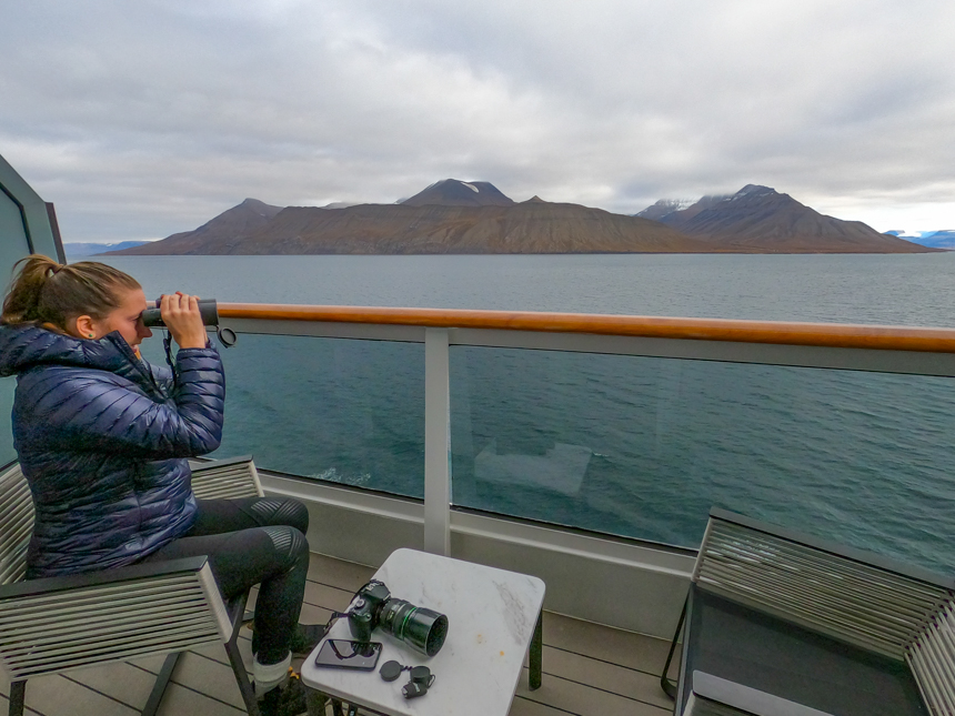 Female in blue jacket sits outside on cabin balcony looking at the landscape with binoculars.
