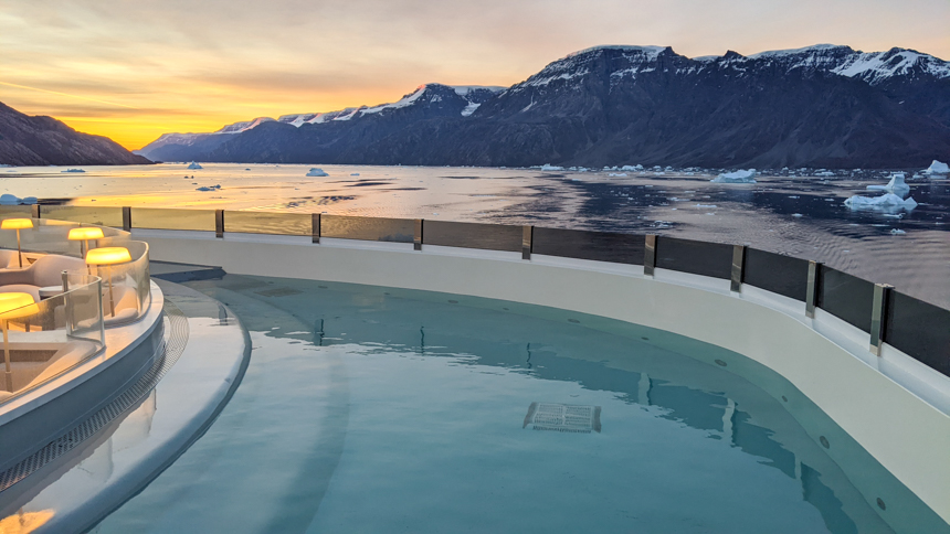 Blue Lagoon pool on the rear of the Charcot ship, teal water with landscape views of mountain range at sunset. 