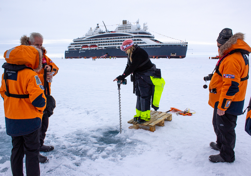 Standing on sea ice surrounded by guests in orange parkas a guide drills into the ice for Citizen Science project. 