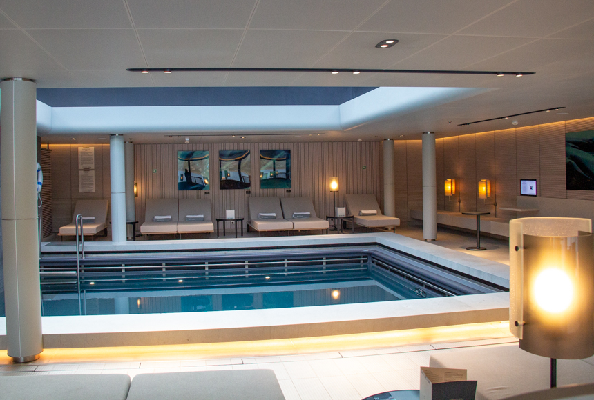 Indoor pool with massive overhead sky light, surrounded by lounge chairs, lamps and artwork by the onboard spa. 