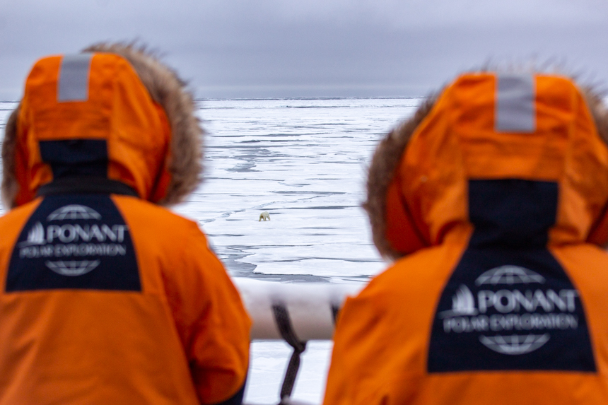 From the ship two travelers in hooded orange parkas watch a polar bear walk along the never ending ice sheet below them. 