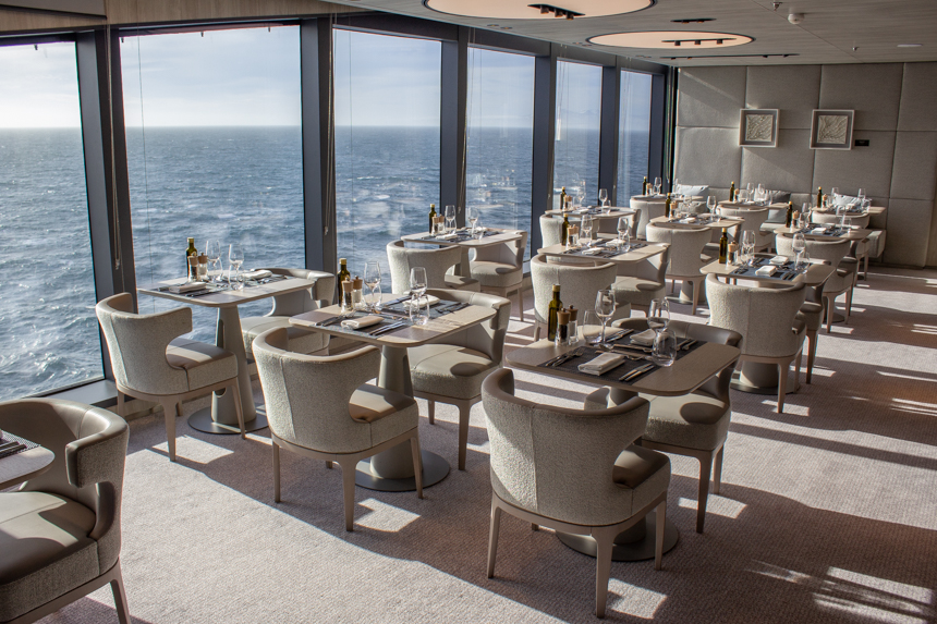 Sila Restaurant Aboard Charcot ship. Small tables with two chairs set along floor to ceiling glass windows with views. 
