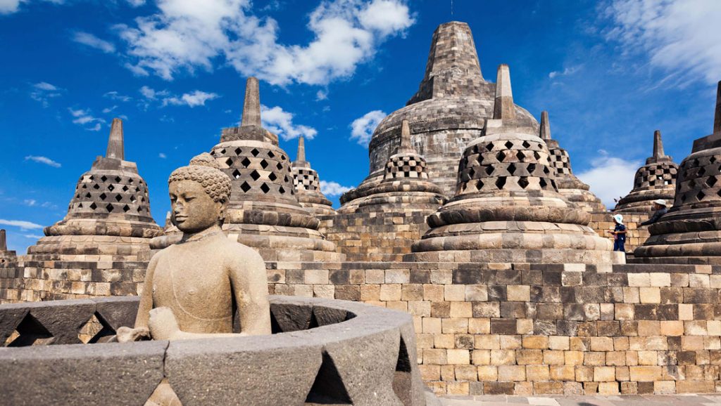 Large stone bell & Buddha statues with varying shades of tan sit atop a stone terrace on a sunny day.
