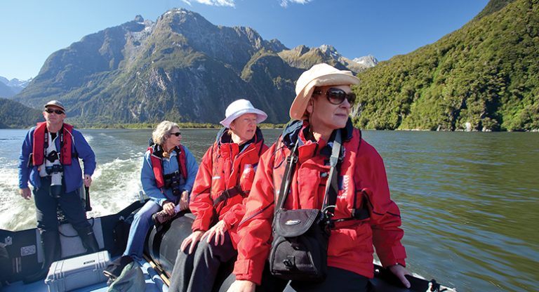 New Zealand travelers on a Zodiac cruise in calm water by green mountains through Fiordland National Park on a sunny day.