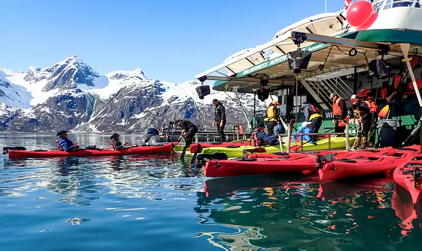 The rear of Wilderness Adventurer ship in Alaska amongst snowcapped mountains. UnCruise crew help guests into red double kayaks.