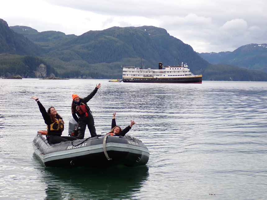 three female uncruise crew members pose on inflatable zodiac as they navigate the water in Alaska around the Wilderness Legacy ship. 