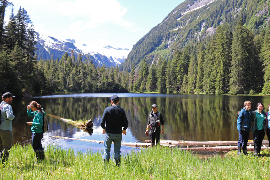 At the end of a bushwhacking activity UnCruise guests stand and take photos in front of an alpine lake surrounded by trees and mountain range. 