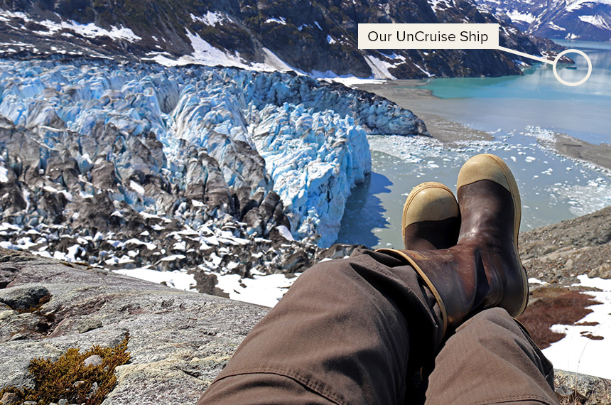 Legs and booted feet shown sitting on a rock overlooking the massive icy blue and white Lamplugh Glacier that opens to an inlet where a small ship floats. 