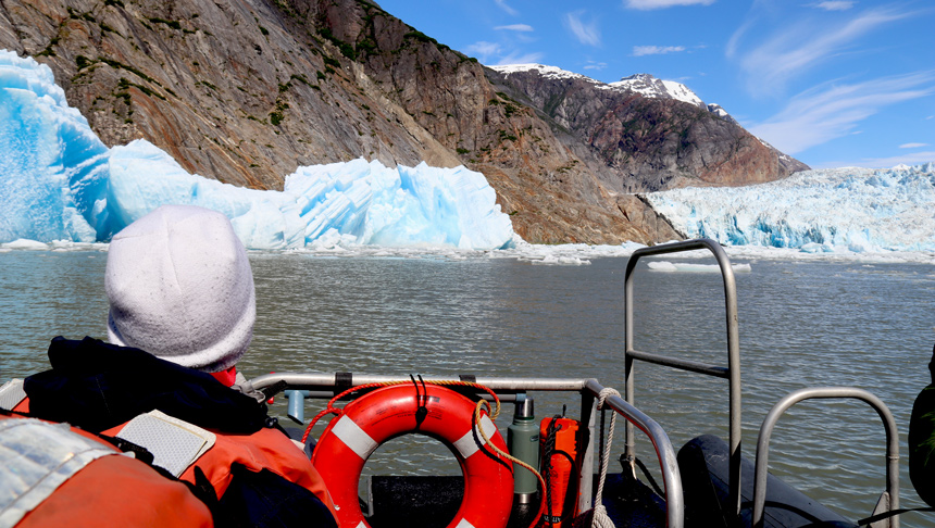 Inflatable skiff navigates a fjord channel in Alaska around ice bergs towards an icy glacier. 