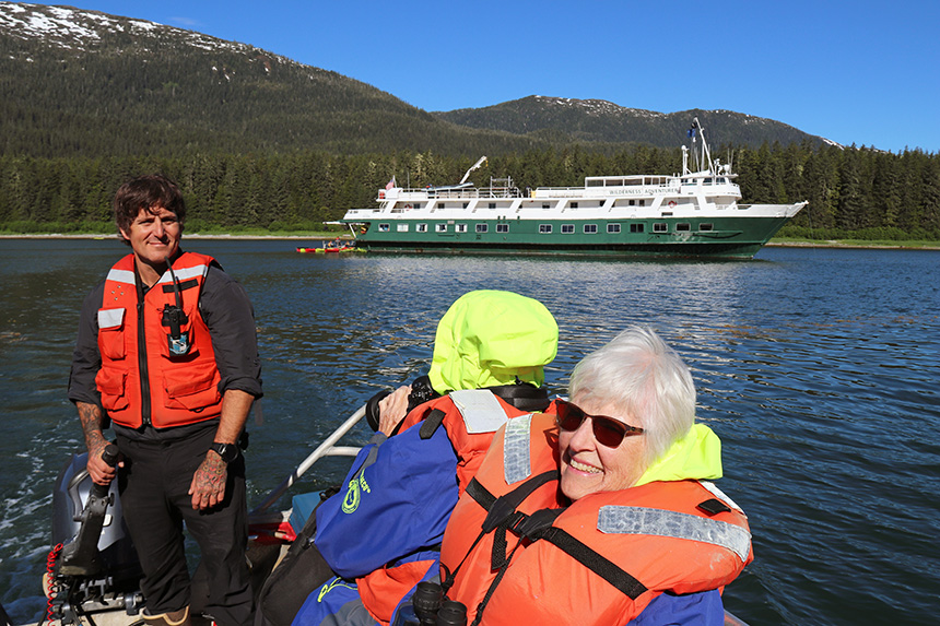 Crew member drives inflatable skiff of cruise guests wearing orange life jackets, away from white and green painted Wilderness Adventurer ship