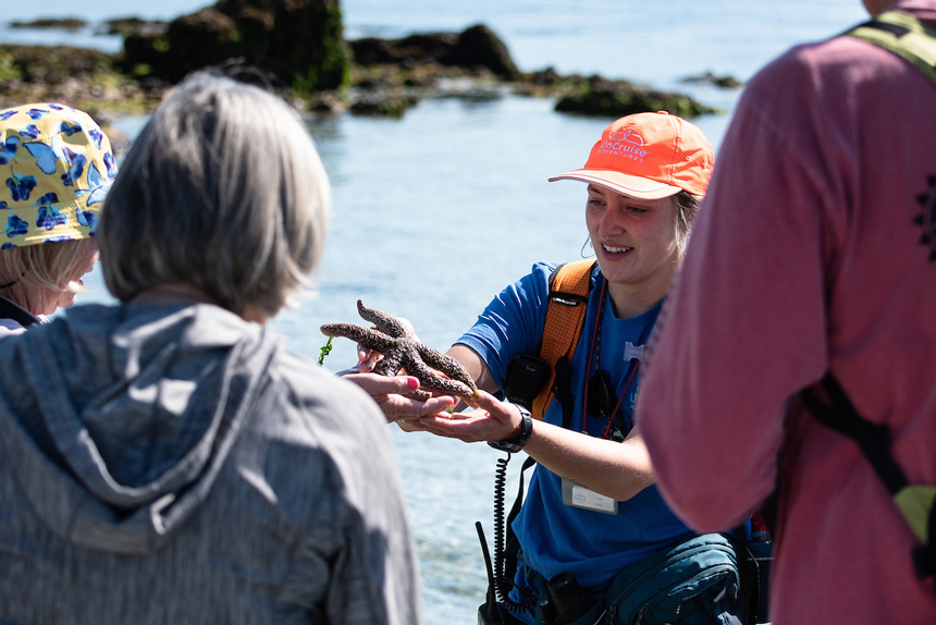 Naturalist guide wearing orange hat with UnCruise logo holds a sea star and speaks to guests during a beachcombing activity.