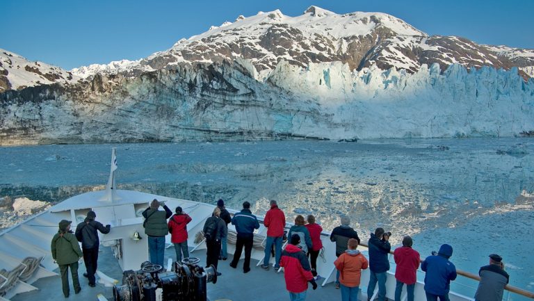 Winter Alaska cruise travelers stand on bow of ship looking at snowy Margerie glacier in fading afternoon light of Glacier Bay.