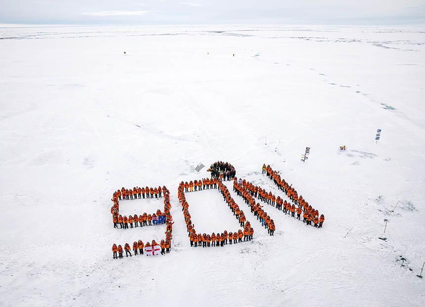 An aerial photo of passengers in orange parkas standing on ice lined up to spell 90N at the North Pole 